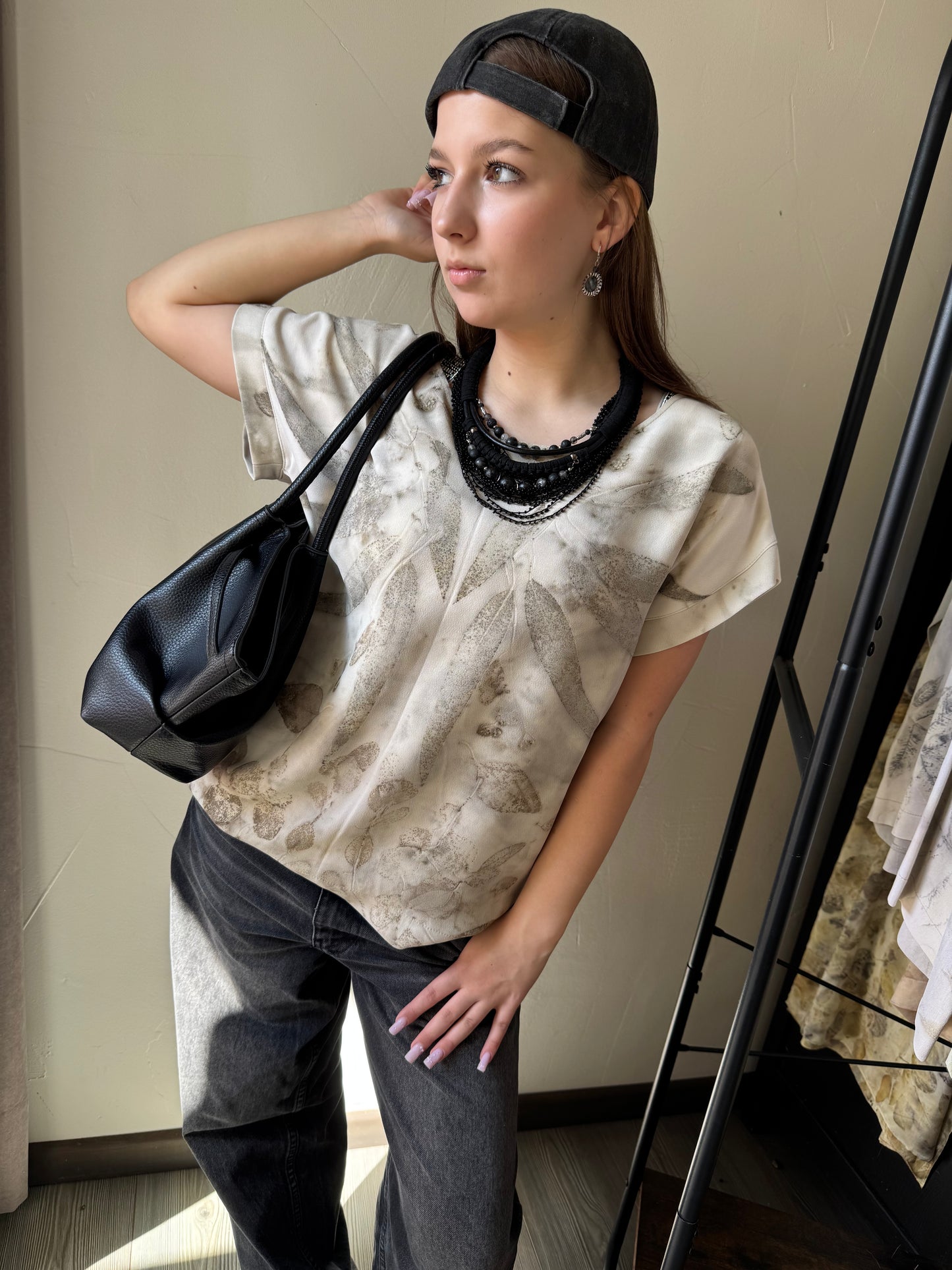 Woman wearing a beige patterned shirt, black pants, and a black cap, holding a black handbag.