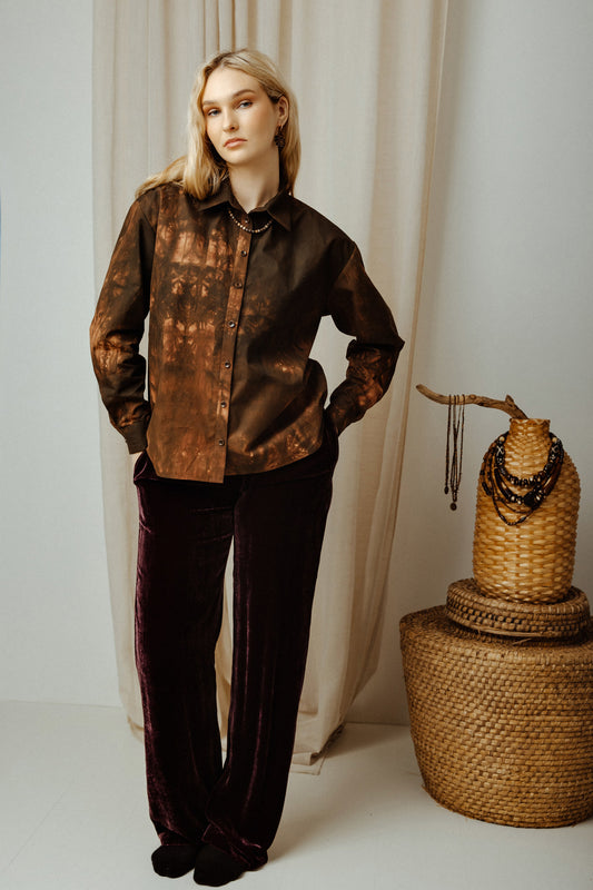 Woman wearing a brown colours tye dye boho shirt and black pants standing next to a woven basket with necklaces.