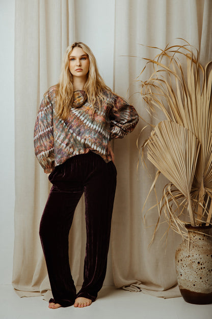 Woman wearing a colourful tye dye blouse with black pants standing against a beige curtain with a plant in the background.