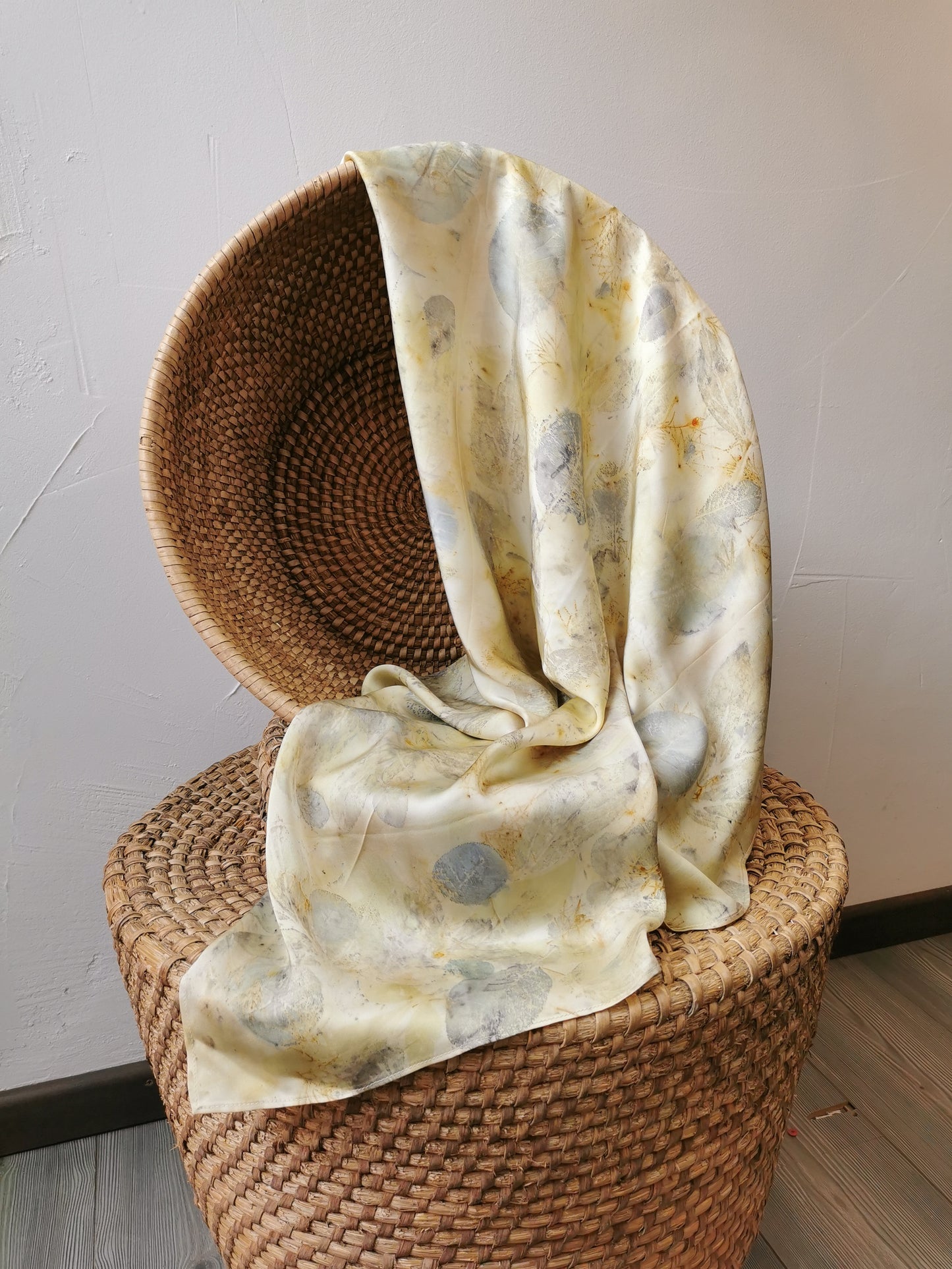 Woven chair with a eco-printed silk scarf draped over it against a plain wall.