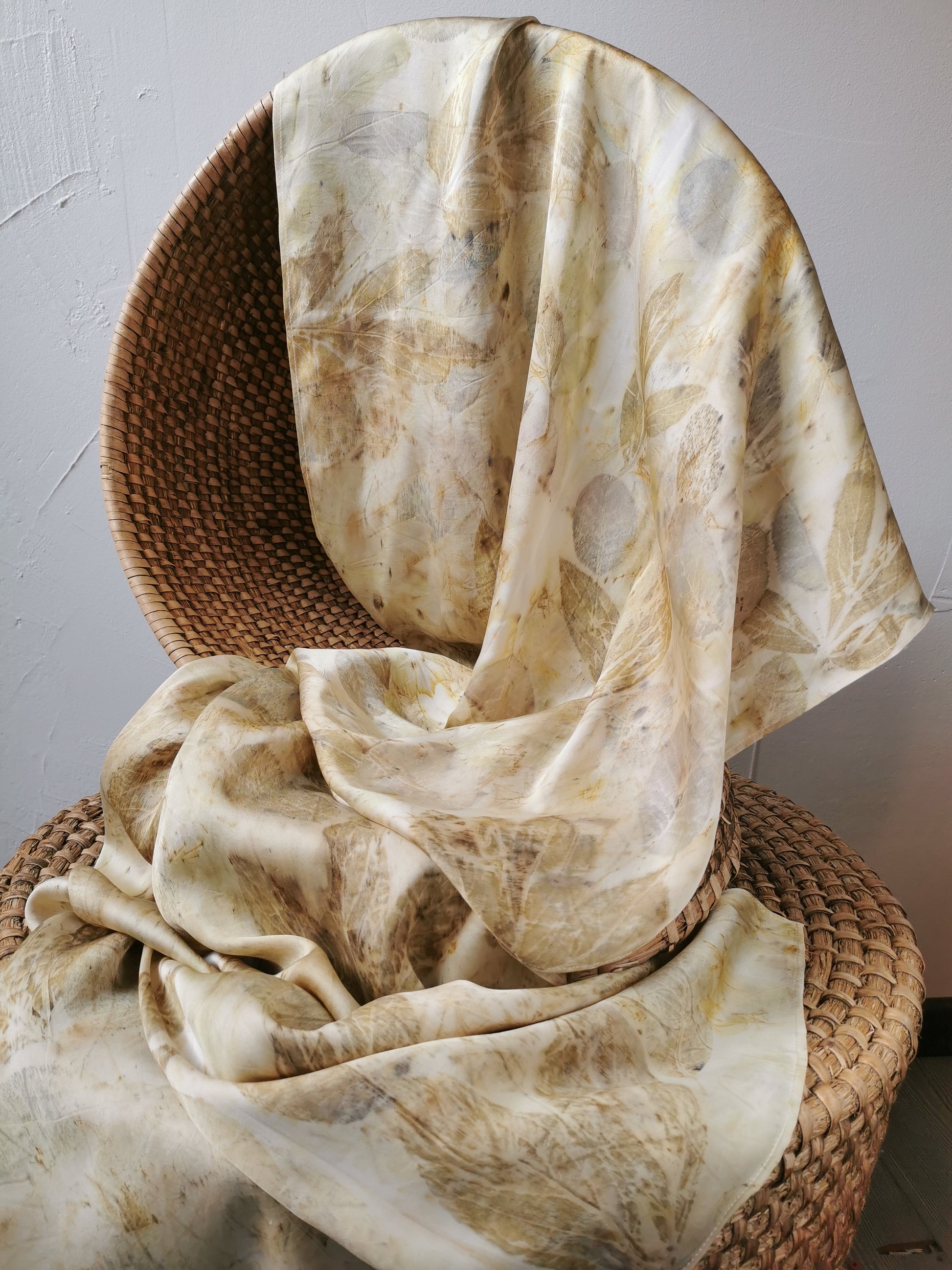 Beige eco-printed silk scarf with floral prints draped over a wicker chair against a plain wall.
