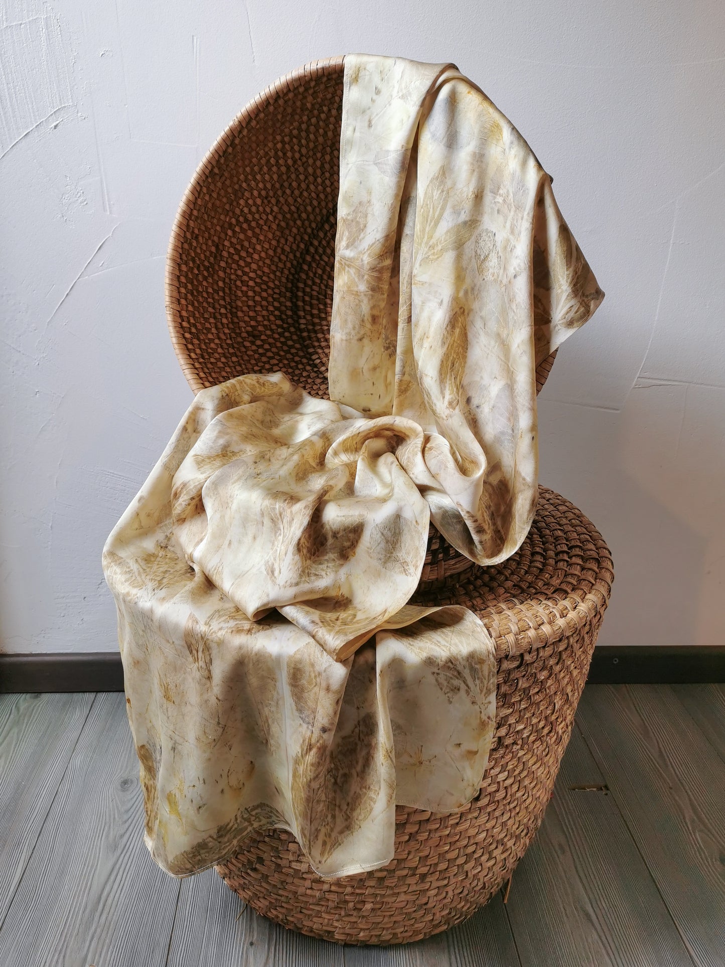 Woven basket with a beige eco-printed silk scarf with floral prints draped over it on a light wooden floor.
