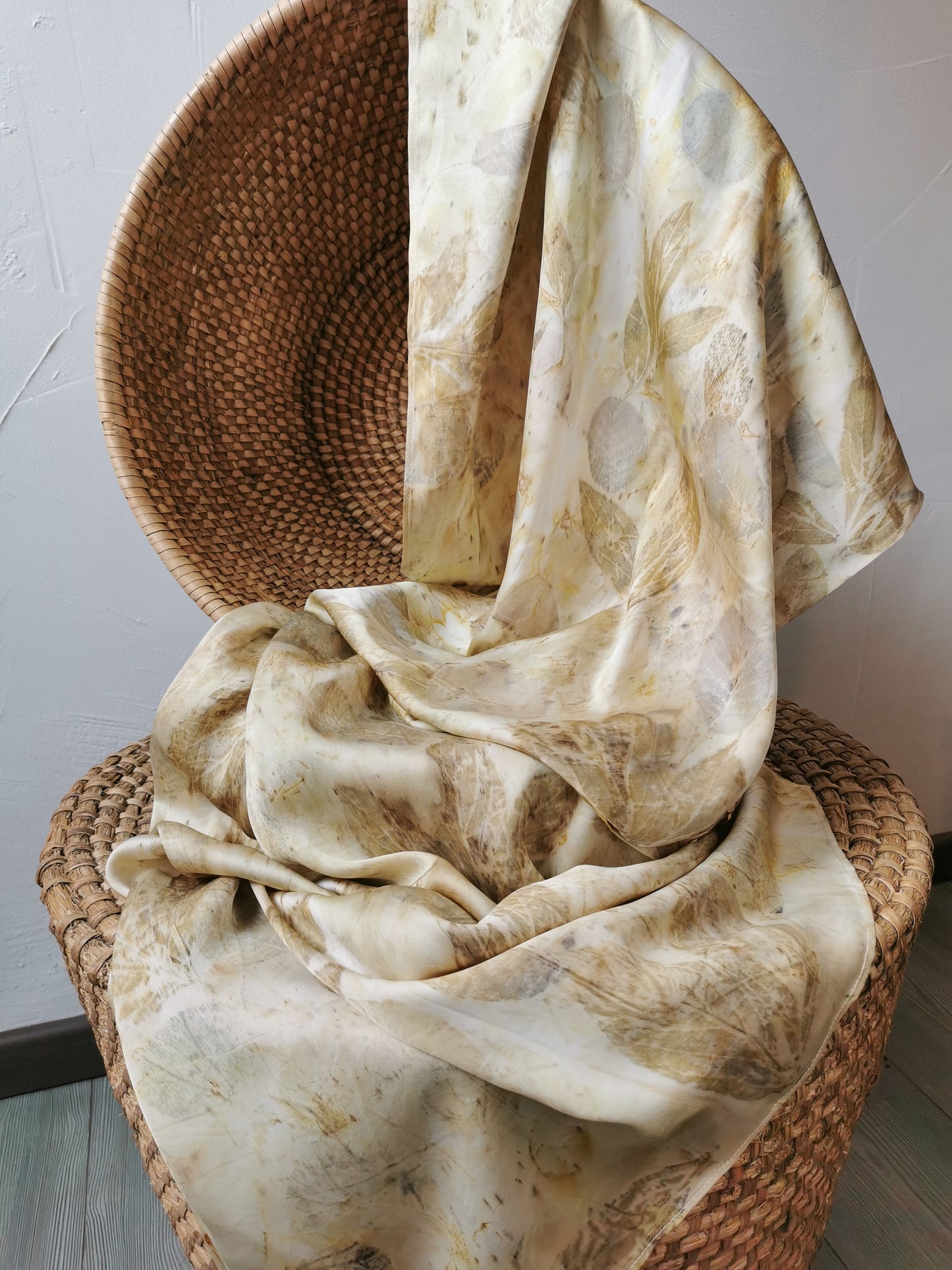 Beige eco-printed silk scarf with floral prints draped over a wicker chair against a neutral background