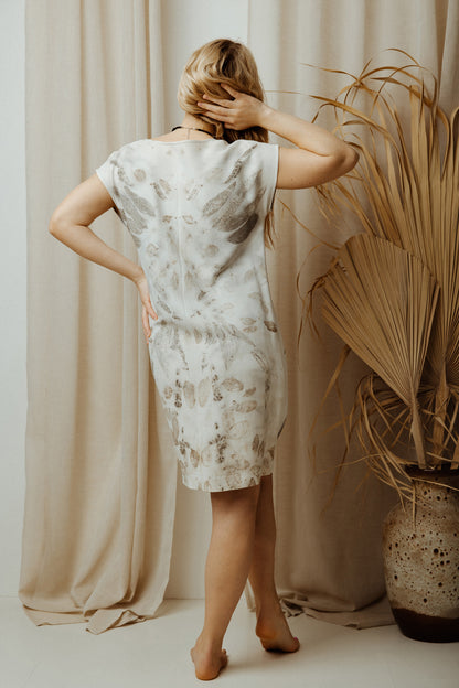 Woman wearing grey eco print viscose dress with floral patterns, standing in a room with beige curtains and a decorative plant.
