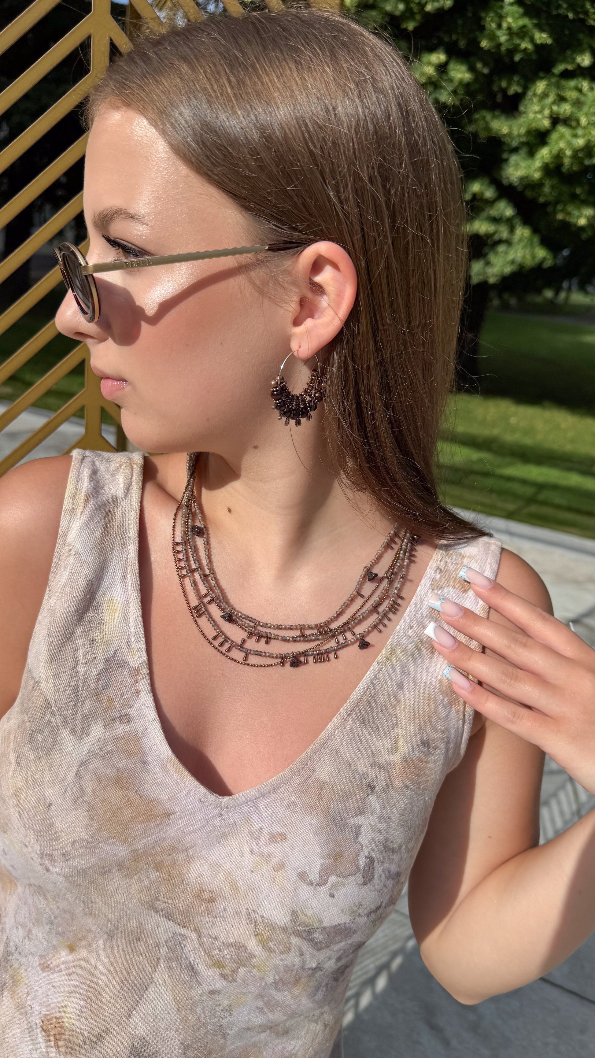 A woman outdoors wearing a layered necklace with dark metallic script lettering, a sleeveless beige patterned top, and sunglasses.