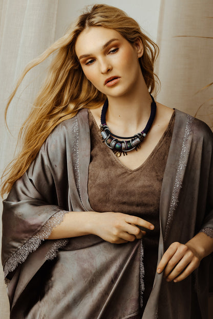 Woman wearing a brown dress with a gray shawl and a blue necklace against a neutral background